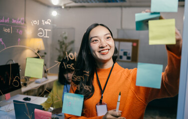 Young manager is smiling while organizing tasks for the day, writing on sticky notes and attaching...