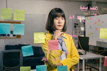 Young professional is thinking about her next move while standing in her office in front of a glass wall covered with sticky notes and project management notes