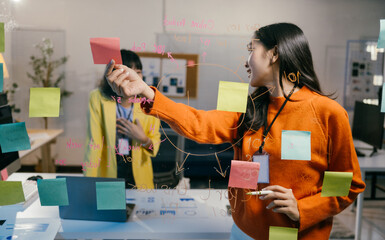 Two businesswomen brainstorm on sticky notes in a dimly lit office at night, focusing on teamwork,...