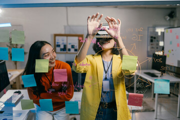 Two young businesswomen are using virtual reality glasses in the office to discuss a project, brainstorming and taking notes on a glass wall
