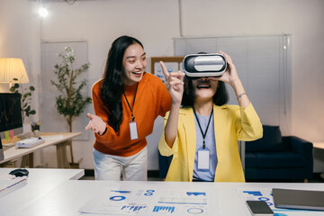 Two businesswomen are having fun using virtual reality glasses in the office while working on a new project. They are looking at graphs and charts on the table