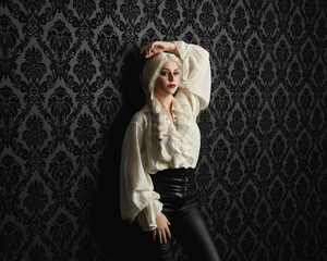 close up portrait of beautiful blonde female model, wearing vintage gothic fantasy ruffle blouse white shirt, leather pants and long blonde plaited hair.  moody black brocade wall paper background