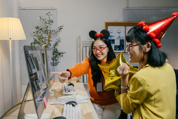 Two young businesswomen are enjoying a successful moment at the office, wearing party hats and holding a balloon while looking at a computer screen