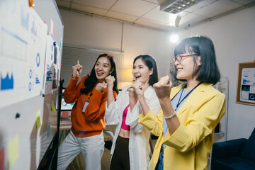 Three asian businesswomen are celebrating success in the office at night, raising their fists in the air and smiling cheerfully