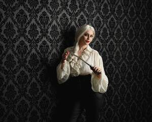 close up portrait of beautiful blonde female model, wearing vintage gothic fantasy ruffle blouse white shirt, leather pants, blonde hair holding knife weapon. moody black brocade wallpaper background