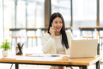 Obraz premium Confident Businesswoman on a Productive Call: Smiling businesswoman in a modern office, using laptop and phone, showcasing efficiency and success. 