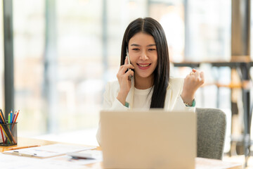 Good News on the Phone: A young Asian businesswoman beams with excitement as she receives positive news on a phone call,  her hand raised in triumph, laptop open in front of her.