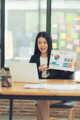 Confident Businesswoman Presenting Data: A young Asian businesswoman confidently presents data charts during a video conference, showcasing professionalism and success in a modern office setting.  