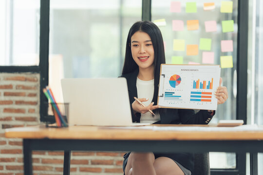 Confident Businesswoman Presenting Data:  A young Asian businesswoman confidently presents financial data during a video conference, showcasing charts and graphs on a clipboard.
