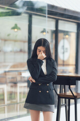 Fototapeta premium A Moment of Reflection: A young woman in a black blazer stands thoughtfully, her expression a mix of contemplation and introspection. The blurred background suggests a busy coffee shop.