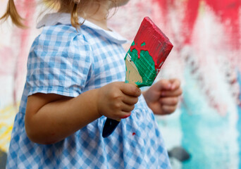 A little girl holding a paintbrush with red and green paint on it