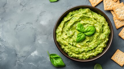 Obraz premium A bowl of creamy guacamole garnished with fresh basil leaves, served with crispy crackers on a textured gray surface.
