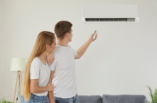 People setting up a comfortable temperature inside the house with air conditioning. Young family couple turning on a modern AC air conditioner on a white wall in a home interior
