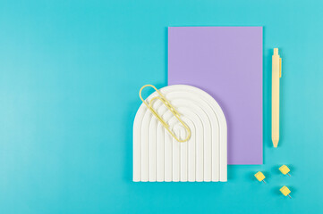 Top view of purple notebook, yellow pen, paperclip, pins, white arch tray on blue background. School, office wallpaper. Flat lay, copy space.