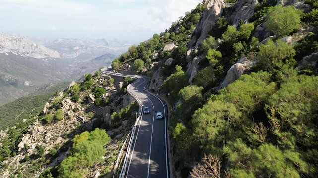 Beautiful aerial shot of a white car driving on the famous road SS 125 in the mountains of Sardinia, Italy