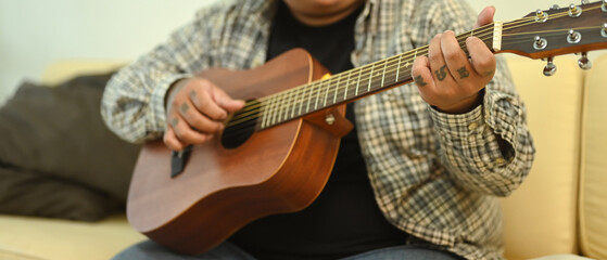 Fototapeta premium Young man playing an acoustic guitar while sitting on sofa at home. Leisure activity and hobby concept