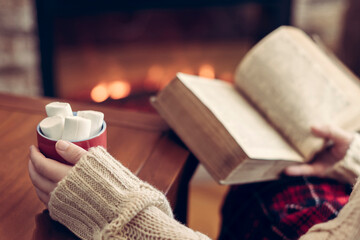 Woman hand with old retro book and cup of hot cocoa and marshmallow on wooden table near christmas...