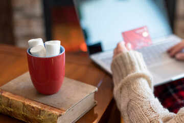 Woman  holding credit card using laptop for making order sitting at table with cup of hot cocoa and marshmallow at christmas fireplace with decoration of light bulbs. Close up.