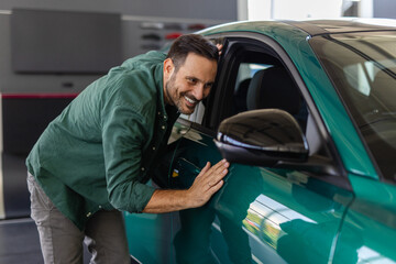 Happy young man hugging his new car in showroom. Satisfied guy with closed eyes embracing the hood...