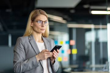 Mature businesswoman in office holding smartphone, embodying leadership and professionalism. Gray suit and glasses highlight focus and confidence in digital communication and business management.