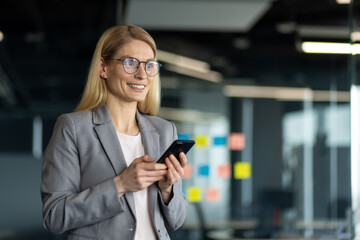 Mature businesswoman with phone in modern office environment exuding confidence and leadership. Her professional attire and demeanor suggest executive role, fostering innovation.