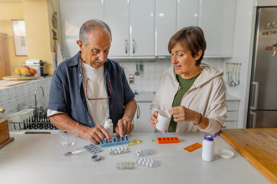 An elderly couple engages in a thoughtful discussion about medication management within a cozy kitchen setting, standing in casual clothing. Organizing pill box, daily routine at home. Horizontal