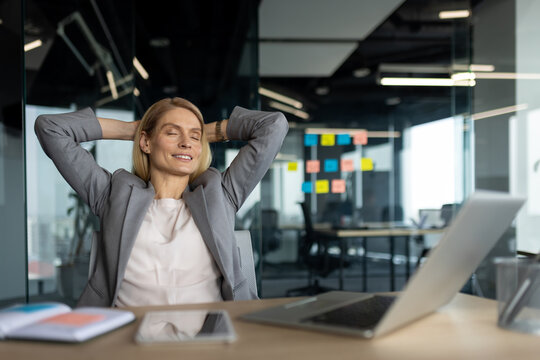 Confident mature business woman takes relaxing break in modern office, leaning back, showing satisfaction. Professional attitude emphasized. Workplace represents corporate success, achievement.