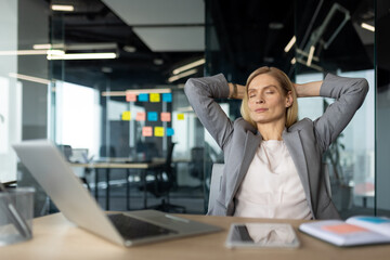 Mature business woman in suit relaxes at desk, embodying focus and calm in busy office setting. She...
