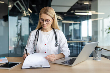 Mature female doctor in professional office setting reviewing medical records. Doctor wearing stethoscope, focused on work at desk with laptop and tablet. Concept: healthcare, professionalism, focus.