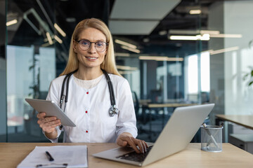 Mature female doctor in office using laptop and tablet for telemedicine. Professional healthcare...