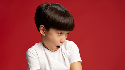 Child, Asian little boy tilting head, mimicking exaggerated shock with wide eyes and open mouth against bold red studio background. Concept of facial-expressions, positive and negative emotions. Ad