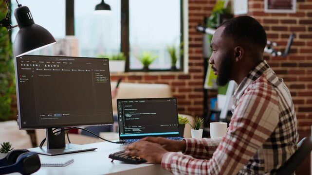 Young man seen programming software code on his laptop in a sleek home office. Workspace with dual monitors helps with software engineering, web development and coding productivity. Camera B.