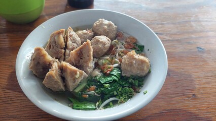 A close-up of a traditional Indonesian meatball soup, known as bakso. The dish consists of savory meatballs served in a light broth with greens, noodles, and fried shallots, offering a comforting, fla