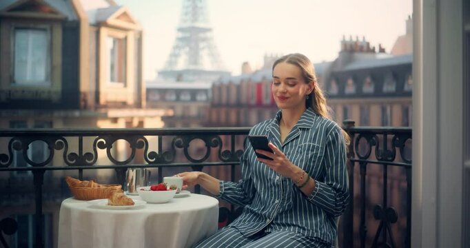 Beautiful Woman in Pajamas Relaxes on a Parisian Balcony, Drinking Coffee and Using Her Smartphone. Eiffel Tower View and Social Media Posts Provide a Perfect Flow to Her Peaceful Morning Routine