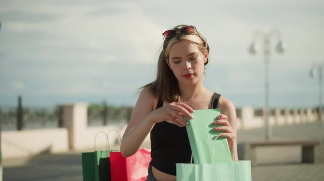 Lady seated on outdoor bench with shopping bags on both sides retrieves a mint color book from mint green bag, opens it, and flips through its pages, while background remains slightly blurred