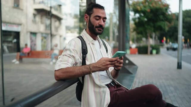 Male tourist using smartphone at bus stop, checking navigation or transport schedule. Dressed casually with backpack, man appears focused, utilizing digital tools for convenient travel planning
