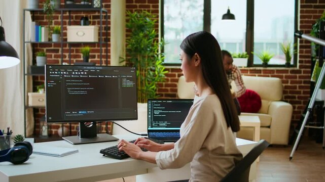 Asian programmer seen typing code for a user interface design on dual monitors, working in a digital workspace. Woman coder working on software engineering and front end development. Camera A.