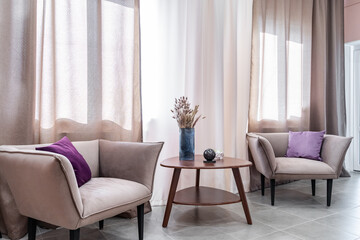 modern interior of living room with soft cozy armchairs, pillows and wooden coffee table. retro floor lamp and brown curtains on windows create a soft warm light. A romantic evening at home for two