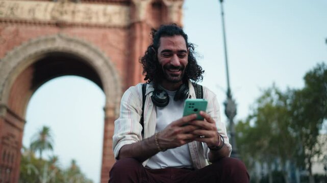 Man smiling while using smartphone in front of Arc de Triomf, enjoying seamless online experience with 5G connectivity. Relaxed and happy male tourist engages in digital communication outdoors
