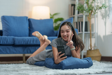 Fototapeta premium Happy young attractive Asian woman lying on carpeted area in living room using mobile phone connected to the internet to play games. online shopping View social media, online chat, technology conce