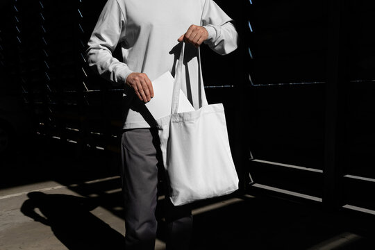 Man pulling a book from the tote bag mockup template in an urban outdoor environment with sharp sunlit shadows. Isolated surface to place your design. 