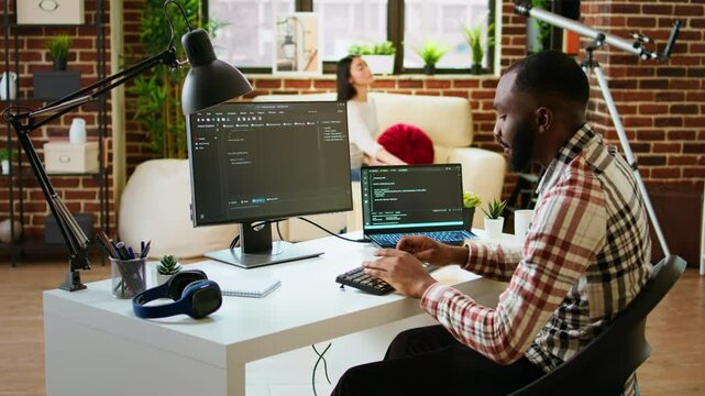 Young man seen programming software code on his laptop in a sleek home office. Workspace with dual monitors helps with software engineering, web development and coding productivity. Camera A.