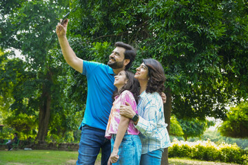 Happy young Indian family of three taking selfie from smart phone at summer park, parents with daughter enjoy vacation together. Love and bonding. Togetherness. Holidays and travel.