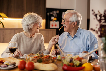 Romantic senior woman is feeding her husband with healthy lunch at dining table at home
