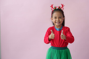 Adorable little Asian girl with Christmas tree headband, antlers showing various poses, standing, smiling, laughing, having fun in various poses, pink background. Holiday concept annual festival.