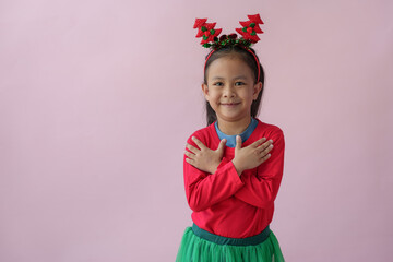 Adorable little Asian girl with Christmas tree headband, antlers showing various poses, standing, smiling, laughing, having fun in various poses, pink background. Holiday concept annual festival.