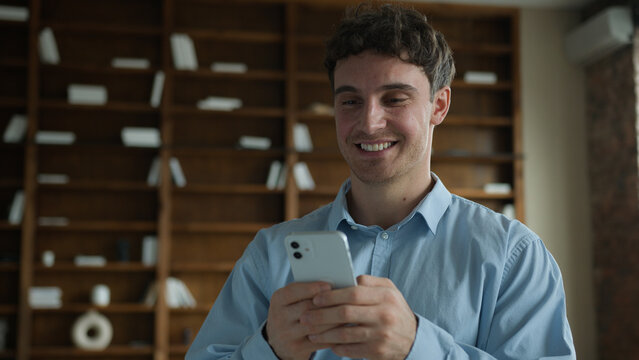 Happy wide smiling young Caucasian male business man businessman guy entrepreneur using mobile phone smartphone typing message chatting browsing internet enjoy online app game ordering delivery indoor