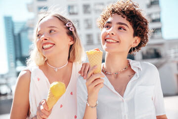 Two young beautiful smiling hipster female in trendy summer clothes. Carefree women posing on street background. Positive models eating tasty ice cream in waffles cone in sunny day, cheerful and happy