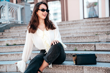 Young beautiful smiling hipster woman in trendy summer white blouse and jeans. Carefree woman posing in the street in sunny day. Positive model outdoors. Cheerful and happy, sits at stairs