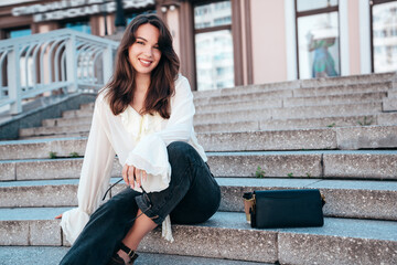Fototapeta premium Young beautiful smiling hipster woman in trendy summer white blouse and jeans. Carefree woman posing in the street in sunny day. Positive model outdoors. Cheerful and happy, sits at stairs